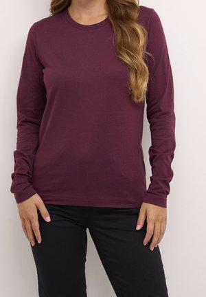 Kobieta ubrana w ciemnofioletową koszulę z długim rękawem i okrągłym dekoltem oraz czarne spodnie, z falującymi jasnobrązowymi włosami opadającymi na jedno ramię.