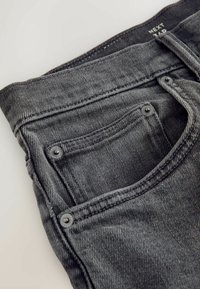Gros plan sur un jean en denim gris foncé montrant la poche avant, la petite poche à monnaie avec rivets, et les coutures visibles.
