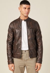 Giacca in finto cuoio marrone con colletto rialzato, due tasche sul petto con zip e una texture liscia. Indossata sopra una camicia bianca e pantaloni beige.