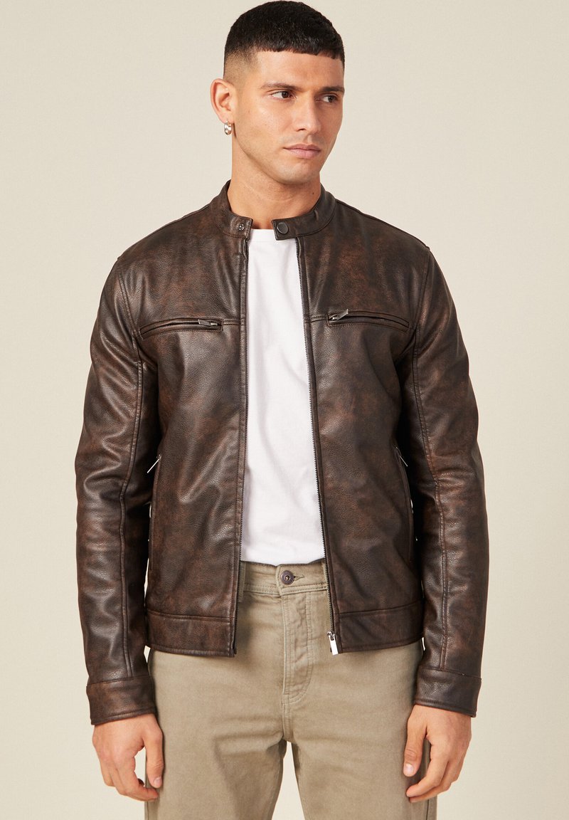 Giacca in finto cuoio marrone con colletto rialzato, due tasche sul petto con zip e una texture liscia. Indossata sopra una camicia bianca e pantaloni beige.