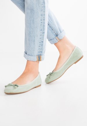 Pieds chaussés de ballerines vert clair avec des nœuds décoratifs, assortis à un jean bleu clair retroussé sur fond blanc.