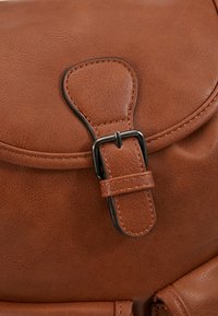 Gros plan sur le rabat d'un sac en cuir marron avec une sangle de boucle cousue et une fermeture à broche métallique.