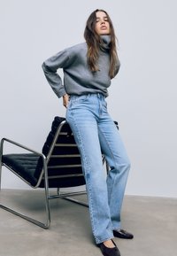 Svetlomodré džínsové nohavice s vysokým pásom a rovnými nohavicami s minimálnym opotrebovaním, skombinované so šedým rolákom, na neutrálnom pozadí.