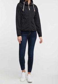 Chaqueta negra con cremallera y capucha, que presenta cordones, dos bolsillos laterales y puños elásticos. Combinada con jeans oscuros y zapatillas blancas.