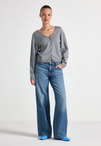 Grijze, knopen-up cardigan met een V-hals, gecombineerd met wijde denim jeans. Blauwe puntige schoenen; soepel materiaal, casual pasvorm.