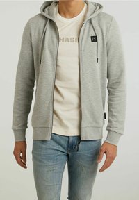 Hombre con sudadera con cremallera de color gris claro sobre camiseta blanca estampada y vaqueros azules desgastados, de pie con los brazos relajados a los lados.