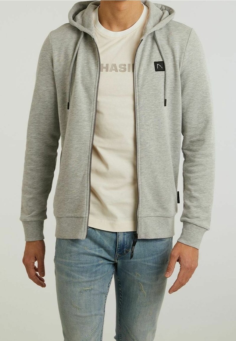 Hombre con sudadera con cremallera de color gris claro sobre camiseta blanca estampada y vaqueros azules desgastados, de pie con los brazos relajados a los lados.