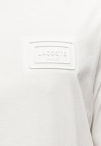 Λευκό βαμβακερό t-shirt με υφασμάτινο λογότυπο από καουτσούκ στην πρόσοψη που γράφει "LACOSTE PARIS". Απλός, καθαρός σχεδιασμός.