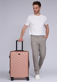 Hartschalenkoffer in Roségold mit Chevron-Muster, schwarzen Akzenten, Teleskopgriff und Rollen für Mobilität. Modell mit hellgrauen Hosen und weißem Hemd.