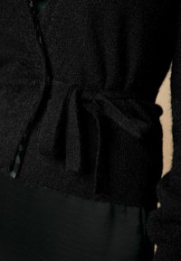 Gilet noir en tricot avec une texture douce, doté d'une taille nouée et d'une coupe légèrement ample, mettant en valeur un design subtil et décontracté.