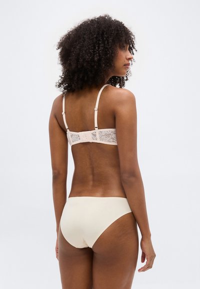 Γυναίκα με σγουρά μαλλιά, φαινομενη από την πίσω πλευρά, φοράει μπεζ δαντελένιο σουτιέν και αντίστοιχο αόρατο εσώρουχο, με φόντο ένα απλό λευκό φόντο.