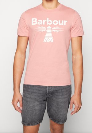 Mężczyzna w różowej koszulce Barbour z białym logo latarni morskiej i ciemnoszarych denimowych szortach na jednolitym tle.