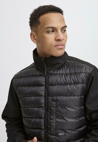 Svart puffervest med høy krage, sydd mønster, glidelås-lukking og sidelomme. Svart stoffermarmer komplimenterer vestens design.