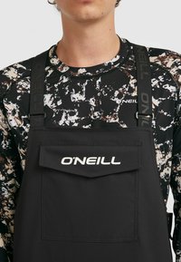 Zwarte O'Neill-pak met een grote voorzak; verstelbare schouderbanden; gedragen over een zwart-wit patroon met lange mouwen.