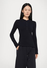 Zwart longsleeve top van zacht materiaal, met een ronde halslijn en een knopenlijst. Draagt bijpassende zwarte broek.