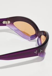 Ochelari de soare mov cu o formă elegantă, având lentile transparente și accente metalice pe tâmple. Textură netedă și design modern.