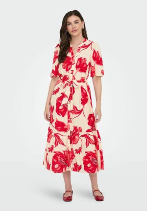 Kvinde iført en cremefarvet midi-kjole med stort rødt blomsterprint, korte ærmer, knaplukning foran, taljebånd og røde flade sko mod en ensfarvet baggrund.