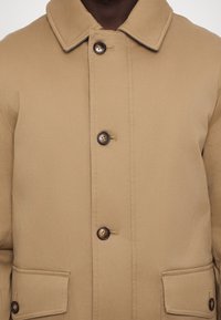 Tan outerwear jacket with a button-up front, large pockets, and a classic collar. Features brown buttons and a smooth fabric texture.