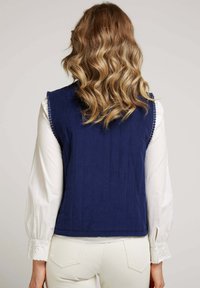 Gilet en velours côtelé bleu marine avec des coutures verticales, un ourlet festonné et des boutons décoratifs sur les épaules, associé à un chemisier blanc.