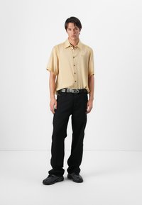 Camisa de manga corta, abotonada en un amarillo pálido, combinada con pantalones negros de corte holgado y zapatos negros. La camisa presenta un logo sutil.