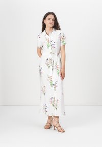 Floral-patterned, short-sleeved dress with a collar, white base, cinched waist, and knee-length hem. Paired with strappy tan heels.