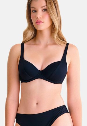 Jeune femme aux longs cheveux blonds portant un soutien-gorge noir uni et une culotte assortie, sur un fond blanc.