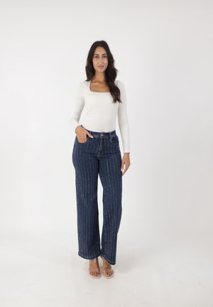 Dunkelblaue Denim-Jeans mit weißen vertikalen Streifen, kombiniert mit einem enganliegenden weißen Langarmoberteil und transparenten Absätzen.