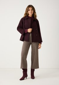 Burgundy wool-blend double-breasted coat with wide sleeves, paired with textured brown plaid trousers and fitted burgundy suede ankle boots.
