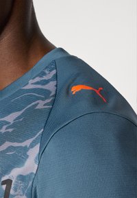 Puma sporto marškiniai su tekstūruota mėlyna medžiaga ir pilku raštu. Oranžinis Puma logotipas siuvinėtas ant peties.