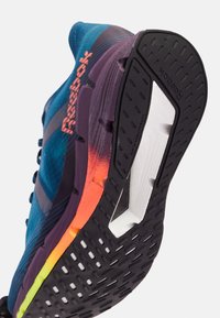Zapatilla de running con suela texturada negra, placa de fibra de carbono visible y parte superior azul con acentos en degradado naranja y amarillo en la mediasuela.
