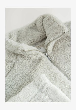 Lys grå zip-up fleecejakke med en blød, tekstureret overflade og høj krave. Har en sølvfarvet lynlås og et mærkat, der er synligt indeni.