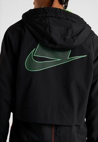 Personne portant une veste à capuche noire Nike avec un grand logo swoosh Nike vert et blanc dans le dos, sur un fond uni.