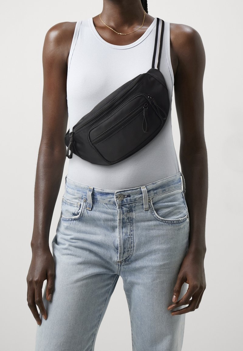 Personne portant un haut blanc sans manches et un jean bleu clair avec un sac bandoulière noir porté en travers de la poitrine.