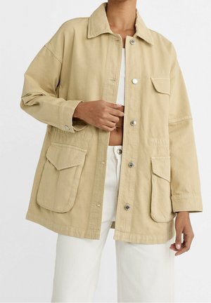 Veste beige oversize avec grandes poches à rabat sur le devant et boutons-pression argentés, portée ouverte sur un top court blanc et un pantalon blanc.