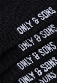 Sorte strikkede sokker med hvid tekst, der siger "ONLY & SONS." Har et enkelt design og blød tekstur. Flere sokker stablet ovenpå hinanden.