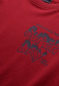 T-shirt in cotone rosso con una stampa grafica nera testurizzata delle parole "SUPER DRY" e "A REAL TEE" sotto di essa. Collo rotondo.