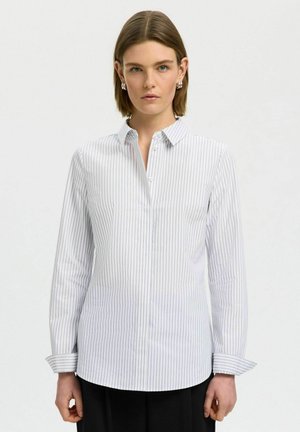 Femme aux cheveux courts et bruns, portant des boucles d’oreilles argentées et une chemise blanche à manches longues avec de fines rayures verticales grises, debout devant un fond uni.