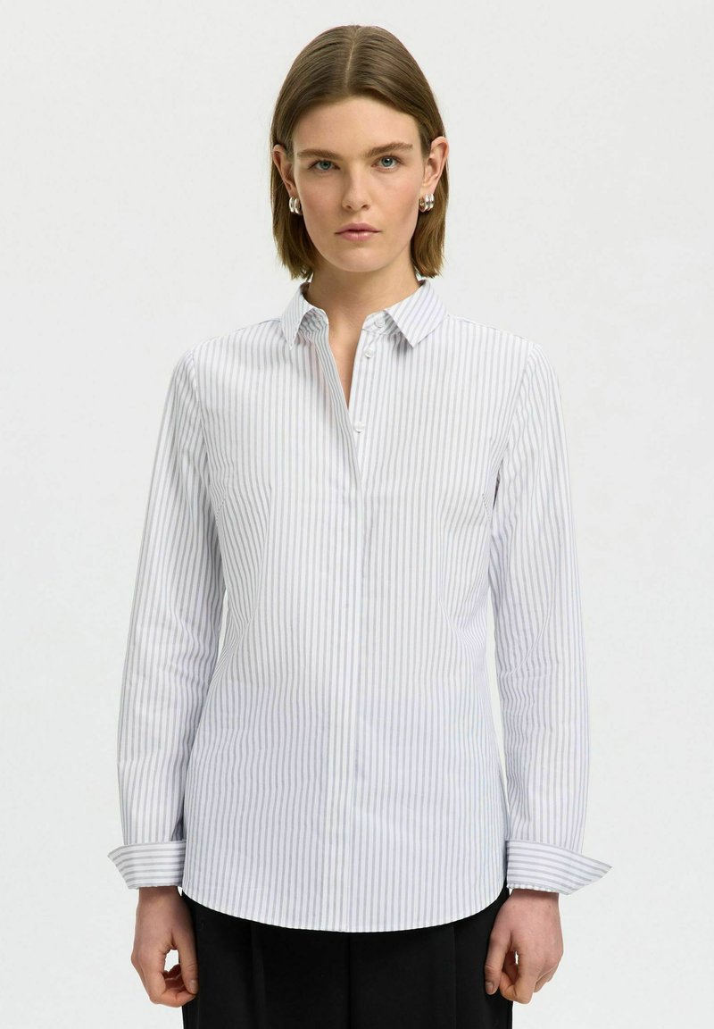 Femme aux cheveux courts et bruns, portant des boucles d’oreilles argentées et une chemise blanche à manches longues avec de fines rayures verticales grises, debout devant un fond uni.