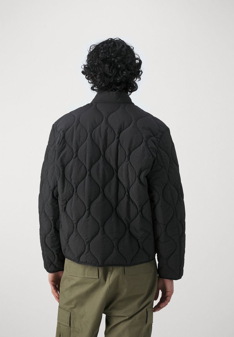 Czarna pikowana kurtka z wysokim kołnierzem, ozdobiona fakturowanym wzorem fal oraz luźnym krojem, zestawiona z oliwkowymi spodniami cargo.