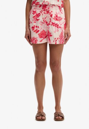Kvinde iført pink og hvide blomstrede shorts med snøre i taljen og brune flade sandaler, stående mod hvid baggrund.
