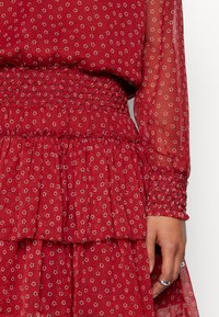 Robe rouge avec un imprimé floral, manches longues transparentes, taille smockée et ourlet superposé à volants. Tissu léger avec une texture douce.