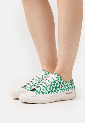 Baskets basses blanches avec des motifs géométriques verts portées pieds nus sur un fond blanc uni.