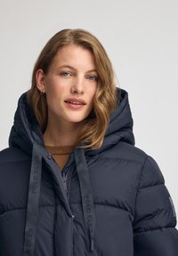 Mörkblå puffjacka med huva, med kviltat mönster, frontdragkedja och märkesdragband. Mjuk textur och avslappnad stil.