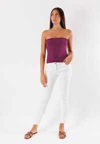 Top senza spalline di colore viola scintillante abbinato a jeans skinny bianchi e sandali piatti marroni; in posizione eretta davanti a uno sfondo neutro.
