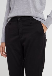 Černé bavlněné kalhoty s rovnými nohavicemi, se zapínáním na knoflík, bočními kapsami a hladkou texturou. Nošeny spolu se šedým svetrem.