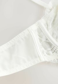 Biely čipkový bralet s priehľadným materiálom, nastaviteľnými ramienkami a jemným lemovaním. Jemný čipkový vzor zvýrazňuje šálky a okraje.