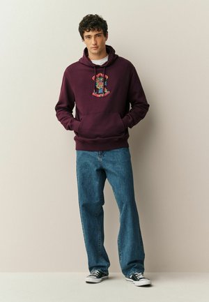 Young man standing with hands in pockets, wearing a maroon hoodie with graphic print, blue jeans, and black sneakers against a plain background.