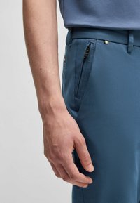 Blue trousers with a smooth texture, featuring a zippered pocket and a sleek waistband, paired with a light blue shirt.