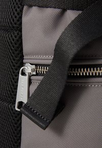 Maille noire et tissu gris avec une fermeture éclair d'un sac. La fermeture éclair est dotée d'une tirette en métal argenté gravée "Carhartt". Surface texturée visible.