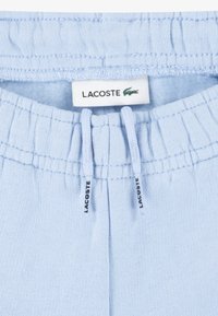 Pantalon de survêtement bleu clair avec une taille élastique, arborant une étiquette Lacoste, des cordons de serrage et une texture de tissu douce.